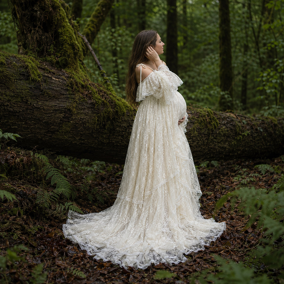 A pregnant woman stands in forest landscape wearing a flowing ivory lace maternity gown with off the shoulder ruffled sleeves and a long, tiered skirt that spreads across the leafy ground.