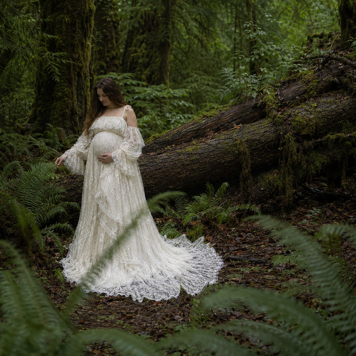 A pregnant woman stands in forest landscape wearing a flowing ivory lace maternity gown with off the shoulder ruffled sleeves and a long, tiered skirt that spreads across the leafy ground.
