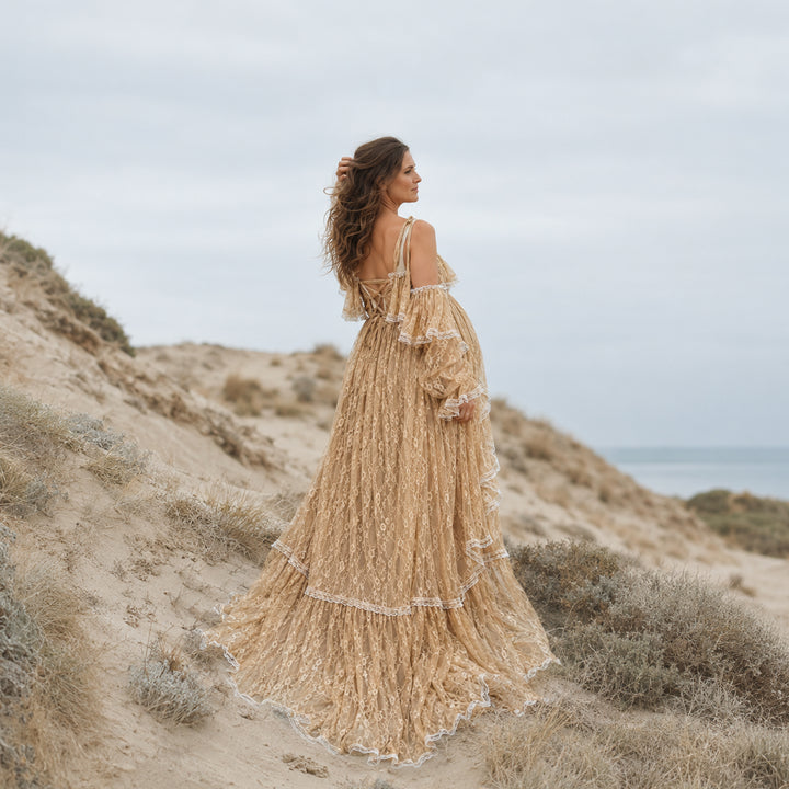 A pregnant woman stands on a sandy coastal dune wearing a flowing khaki melinoe lace maternity gown with tiered ruffles, bell sleeves, and an open back with crisscross straps.