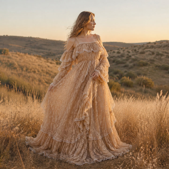 A pregnant woman stands in a sunlit, dry grassy landscape wearing a flowing lace maternity gown with off-the-shoulder ruffled sleeves and a tiered full skirt that spreads across the ground.