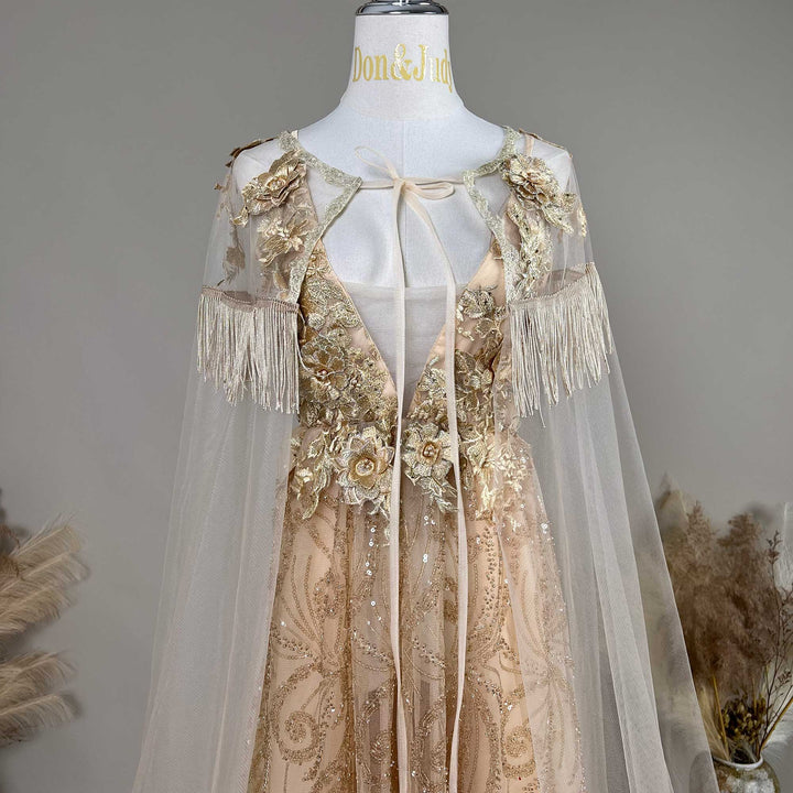 A mannequin displays a gold sequined gown with a tulle cape, featuring 3D floral decorations and fringe cuffs.