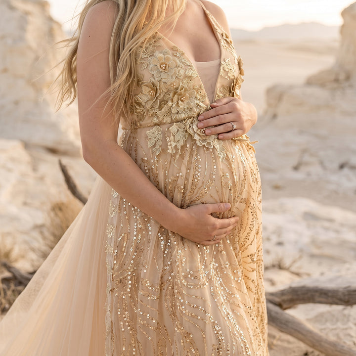 A pregnant model in a gold sequined spaghetti strap gown holds her belly; the dress features 3D floral embroidery and sequins, with desert rocks in the background.
