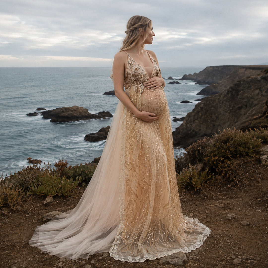 A pregnant model in a gold sequined spaghetti strap gown stands on a seaside cliff, hands on belly.