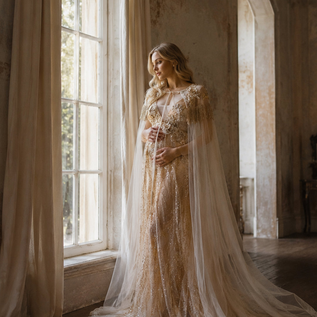 A pregnant model in a Pandia gold sequined gown with a tulle cape stands by a window, hands on belly.
