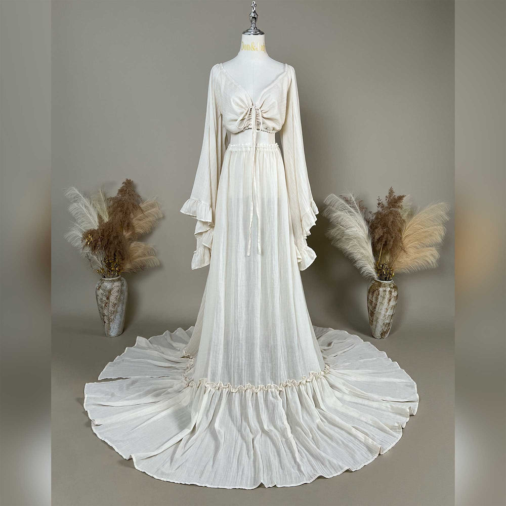 A natural off white two-piece gown is displayed on a mannequin; the top is a deep-V boho crop with tiered trumpet sleeves, and the skirt is a high-waisted floor-length style with a ruffled hem.