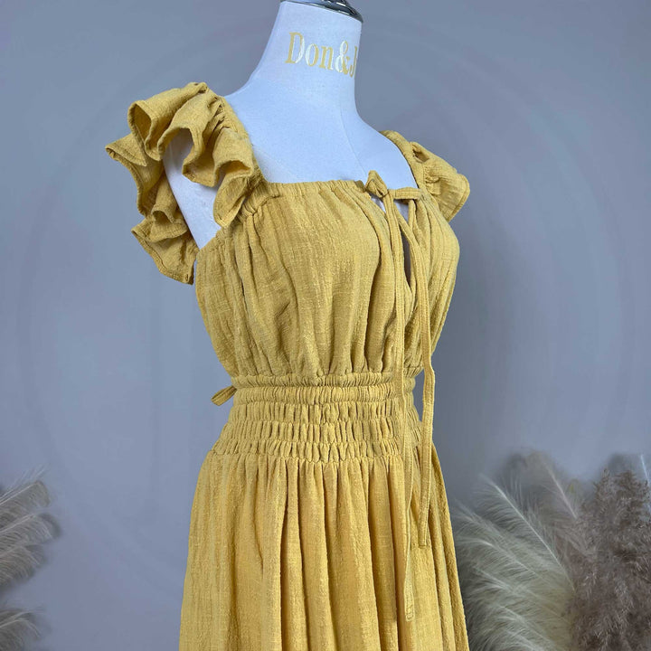 A Tethys gown is displayed on a mannequin, featuring ruffled short sleeves, a front tie, and an elasticated waist; dried flowers are placed on both sides.