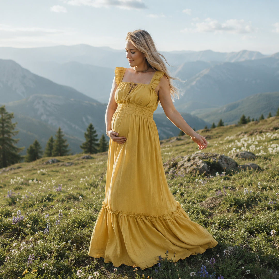 A pregnant woman in a yellow square-neck gown stands on a mountain meadow; the dress has ruffled straps, a front tie, elasticated waist, and tiered ruffled hem. 