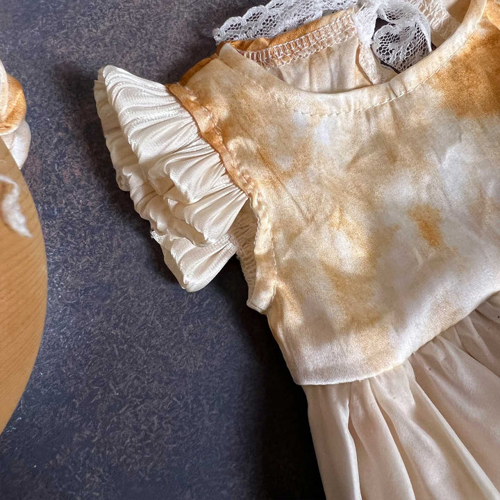 A close-up of a yellow tie-dye baby romper dress, showing the round neck, white lace and one ruffled sleeve, laid on a dark surface.