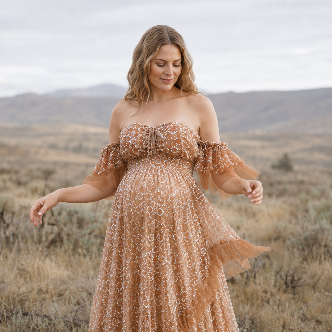 A pregnant woman in a brown sheer off-shoulder gown with white embroidery; she looks down with hands hanging naturally.