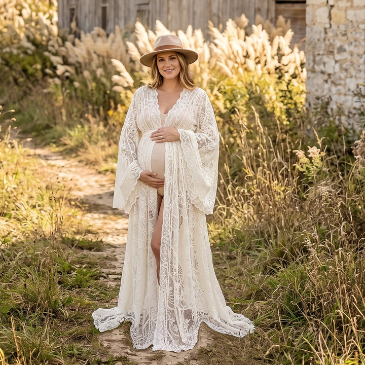 A pregnant woman stands outdoors in front of a wooden structure wearing a beige lace maternity robe with scalloped edges and bell sleeves, with a wide-brim hat.