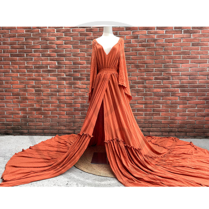 A Daphne rust red long-sleeved V-neck gown is displayed on a mannequin. The dress features a high front slit and its hem and train are spread out on the wooden floor.