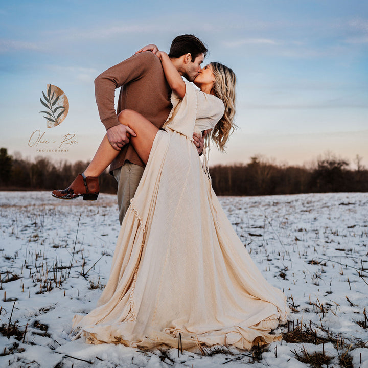 A man lifts a woman in a Daphne long dress in a snowy field while they kiss.