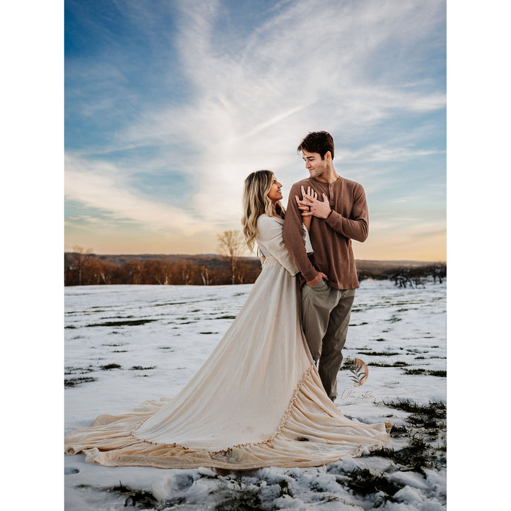 A woman in a beige Daphne long dress and a man in a brown top stand facing each other on snow. A watermark appears at the bottom right.