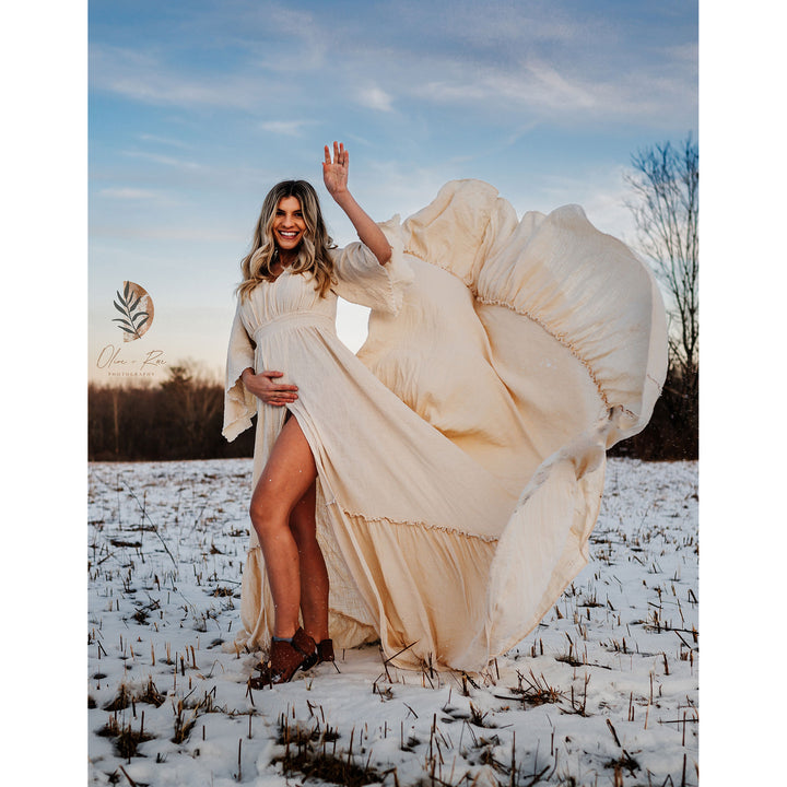 A pregnant woman in a Daphne beige side-slit dress stands in the snow, holding her belly. Her skirt flies up, one hand raised.